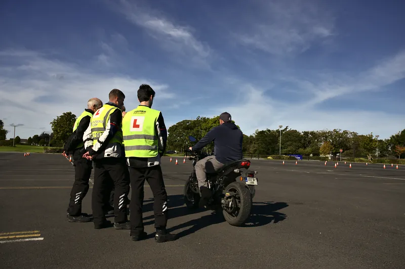 Learners in L-plate vests watching an instructor on a motorcycle at the training yard